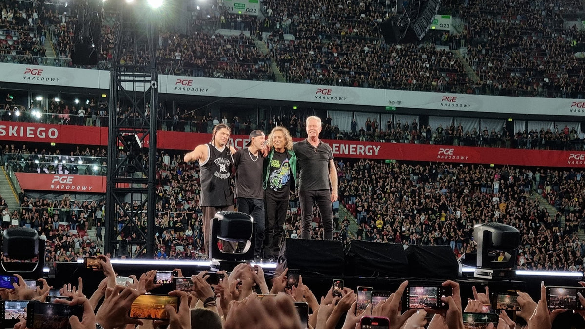 Metallica performing live on stage at a stadium concert in Warsaw, with the band together at the front of the stage and fans holding up phones